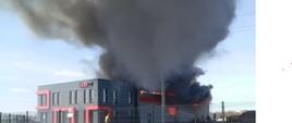 Pożar hali magazynowej. Ponad dach budynku wychodzą płomienie. Nad budynkiem kłęby dymu