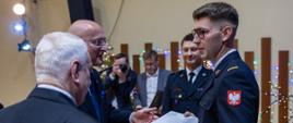 dwie osoby w garniturach wręcza strażakowi list obok stoi inny funkcjonariusz, w tle fotograf oraz osoba prowadząca uroczystość