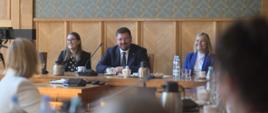 Za drewnianym stołem siedzi wiceminister Rzymkowski, obok niego ubrana na niebiesko kobieta, z drugiej strony kobieta w czarnym ubraniu.