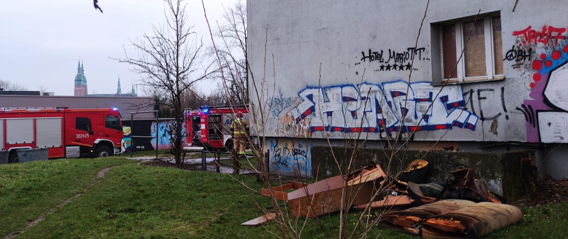Zdjęcie przedstawia budynek socjalny oraz leżące przy ścianie meble zniszczone przez pożar. W głębi widać samochody pożarnicze