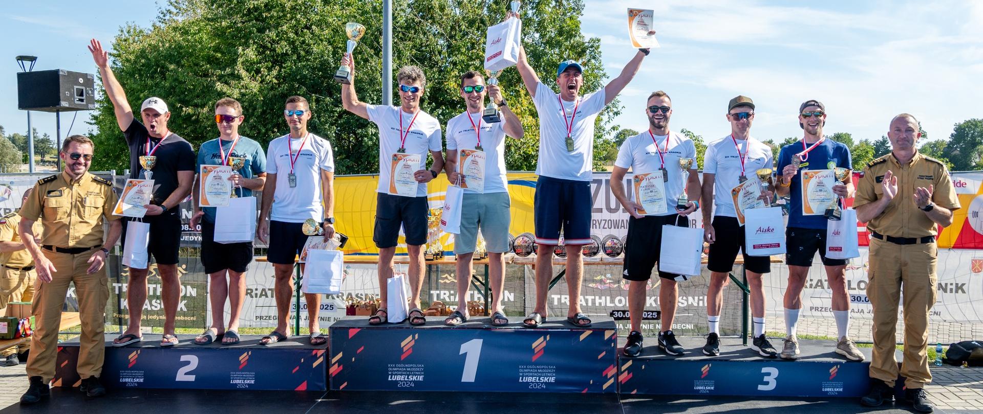 Grupa zawodników na podium podczas ceremonii VI Mistrzostw Polski Strażaków w Triathlonie. Triumfatorzy stoją z medalami, pucharami i dyplomami, unosząc ręce w geście zwycięstwa, obok nich strażacy PSP w mundurach.