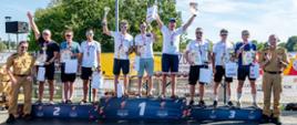 Grupa zawodników na podium podczas ceremonii VI Mistrzostw Polski Strażaków w Triathlonie. Triumfatorzy stoją z medalami, pucharami i dyplomami, unosząc ręce w geście zwycięstwa, obok nich strażacy PSP w mundurach.