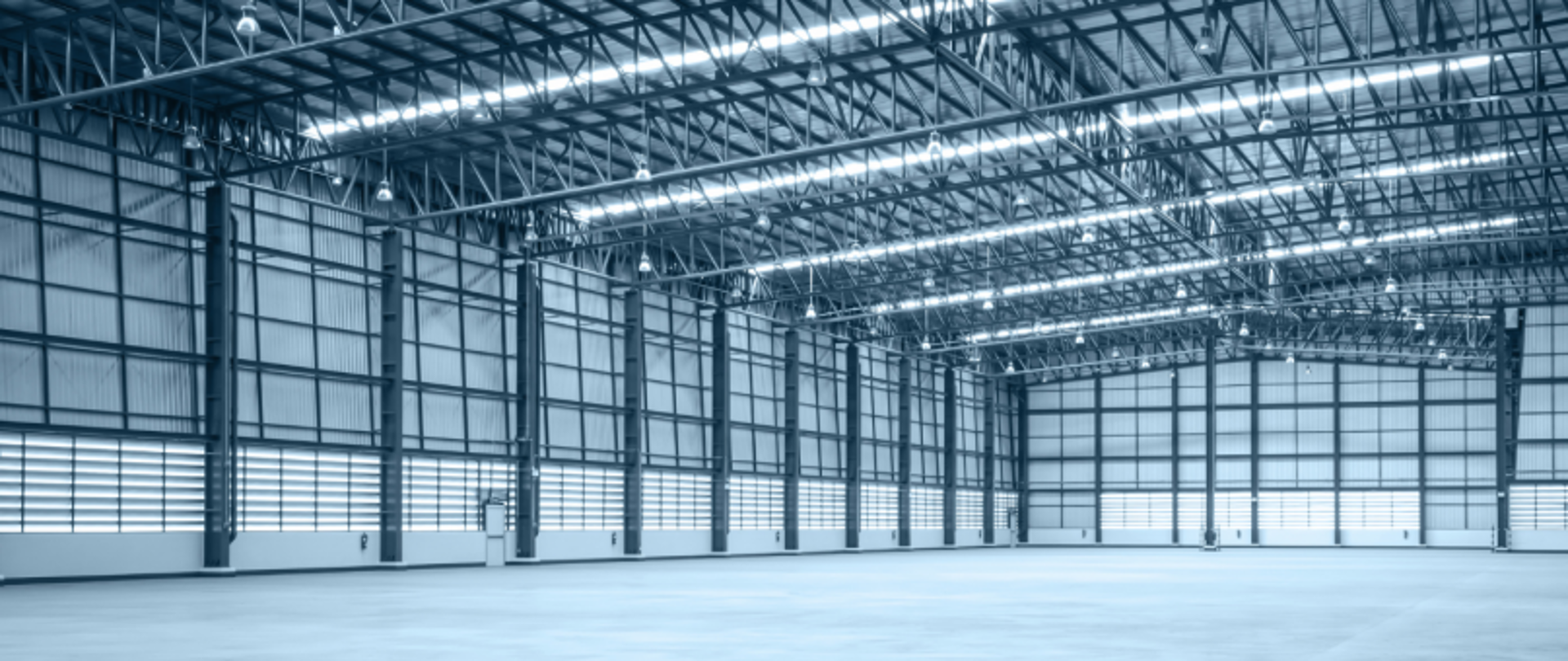 Wnętrze dużego, pustego magazynu z wysokim sufitem, metalowymi ścianami i betonową podłogą, oświetlonego światłem dziennym i lampami sufitowymi.