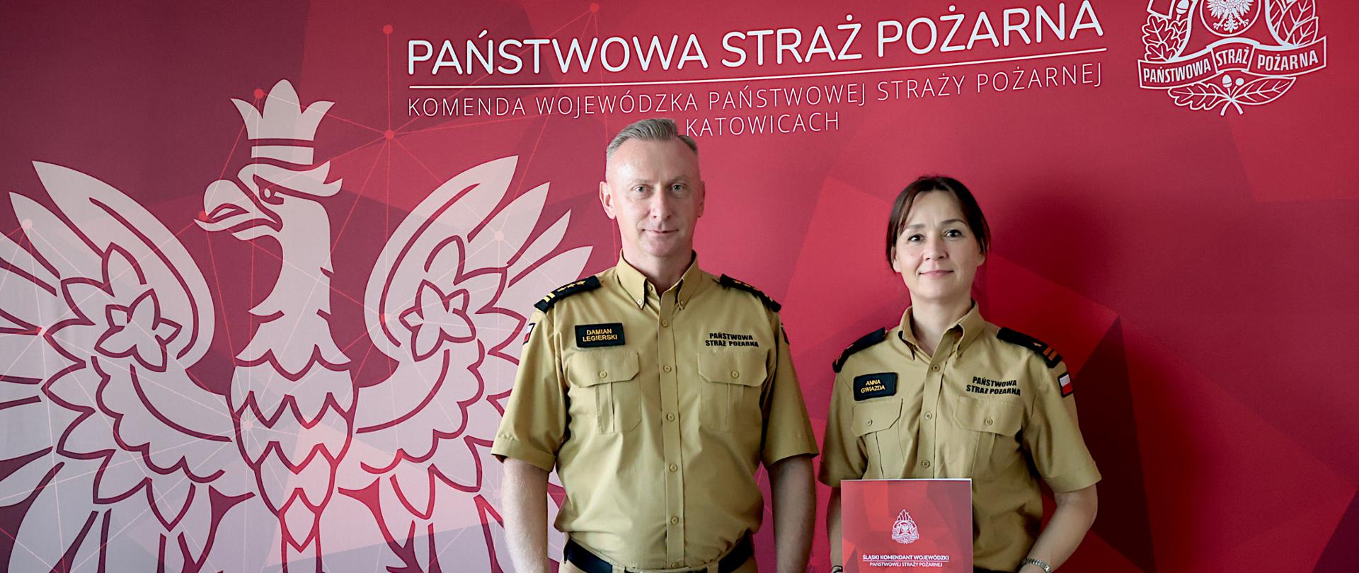 Na zdjęciu stoją obok siebie dwie osoby w piaskowym umundurowaniu służbowym Państwowej Straży Pożarnej. W centrum znajduje się mianowana na zastępcę naczelnika wydziału kadr funkcjonariuszka mł. bryg. Anna Gwiazda, która trzyma w obu dłoniach czerwoną teczkę z godłem PSP i napisem „Śląski Komendant Wojewódzki Państwowej Straży Pożarnej”. Po jej lewej stronie stoi mężczyzna w mundurze służbowym - Śląski komendant wojewódzki Państwowej Straży Pożarnej - st. bryg. Damian Legierski. Wszystkie osoby zwrócone są przodem do obiektywu, stoją wyprostowane i lekko się uśmiechają.
Tłem fotografii jest czerwona ścianka z dużym, białym konturem orła z godła państwowego oraz napisami: „Państwowa Straż Pożarna” i „Komenda Wojewódzka Państwowej Straży Pożarnej w Katowicach”. W prawym górnym rogu widoczne jest logo Państwowej Straży Pożarnej. 