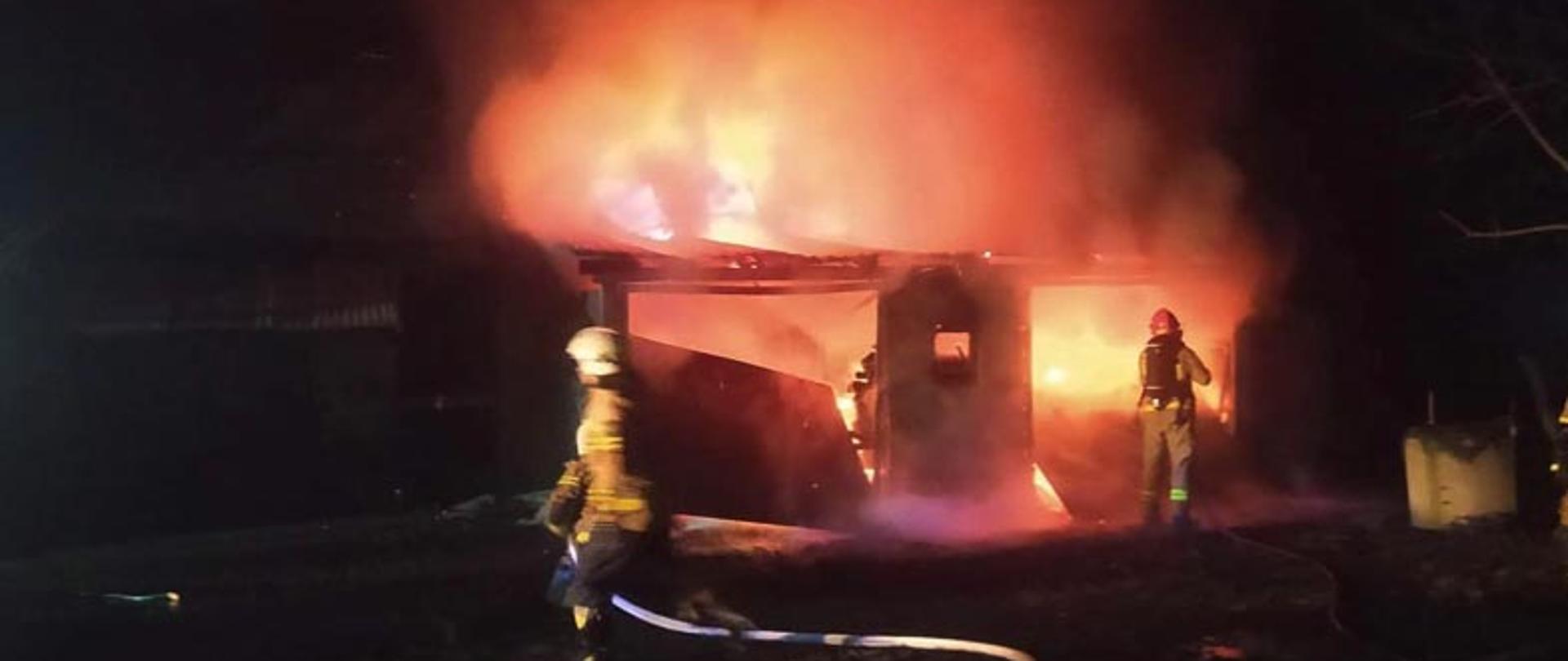 wewnątrz budynku gospodarczego palący się samochód , widoczne płomienie oraz dym przed budynkiem rozwinięta linia wężowa oraz dwóch strażaków 