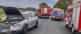 Zdjęcie przedstawia rozbity pojazd koloru szarego. Na drodze stoją również pojazdy straży pożarnej oraz karetka pogotowia