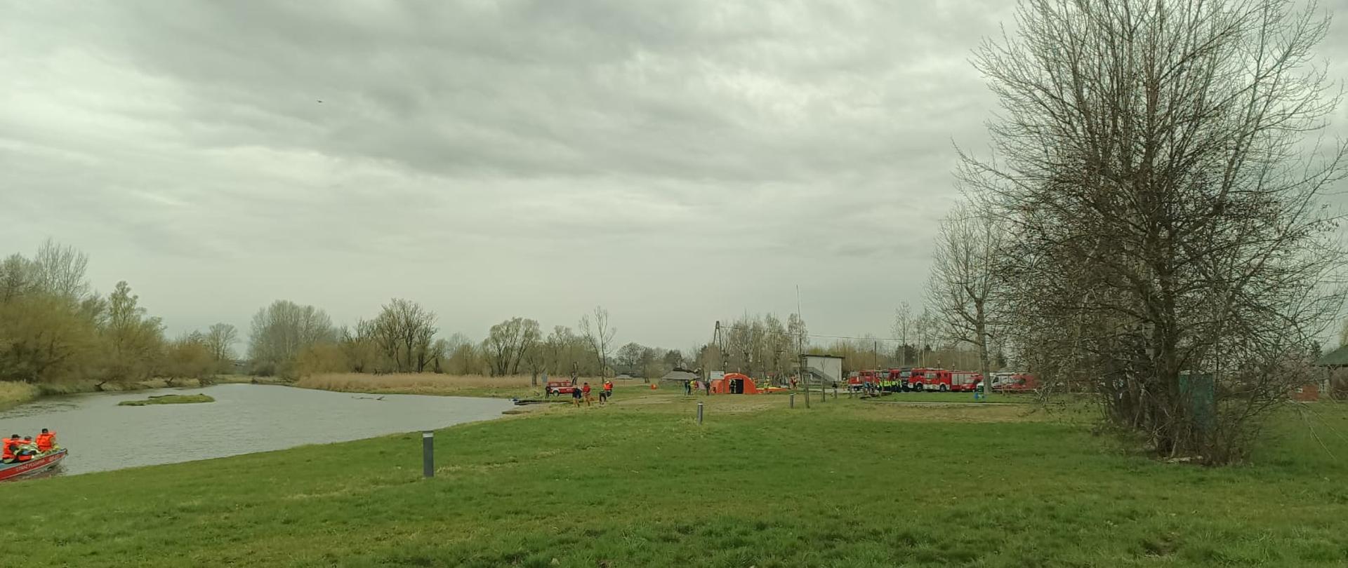 Fotografia przedstawia szeroki widok na rzekę Nidę i miejsce koncentracji sił i środków: widać stojące samochody, namiot, przemieszczających się ratowników a także płynącą rzeką łódkę ze strażakami.