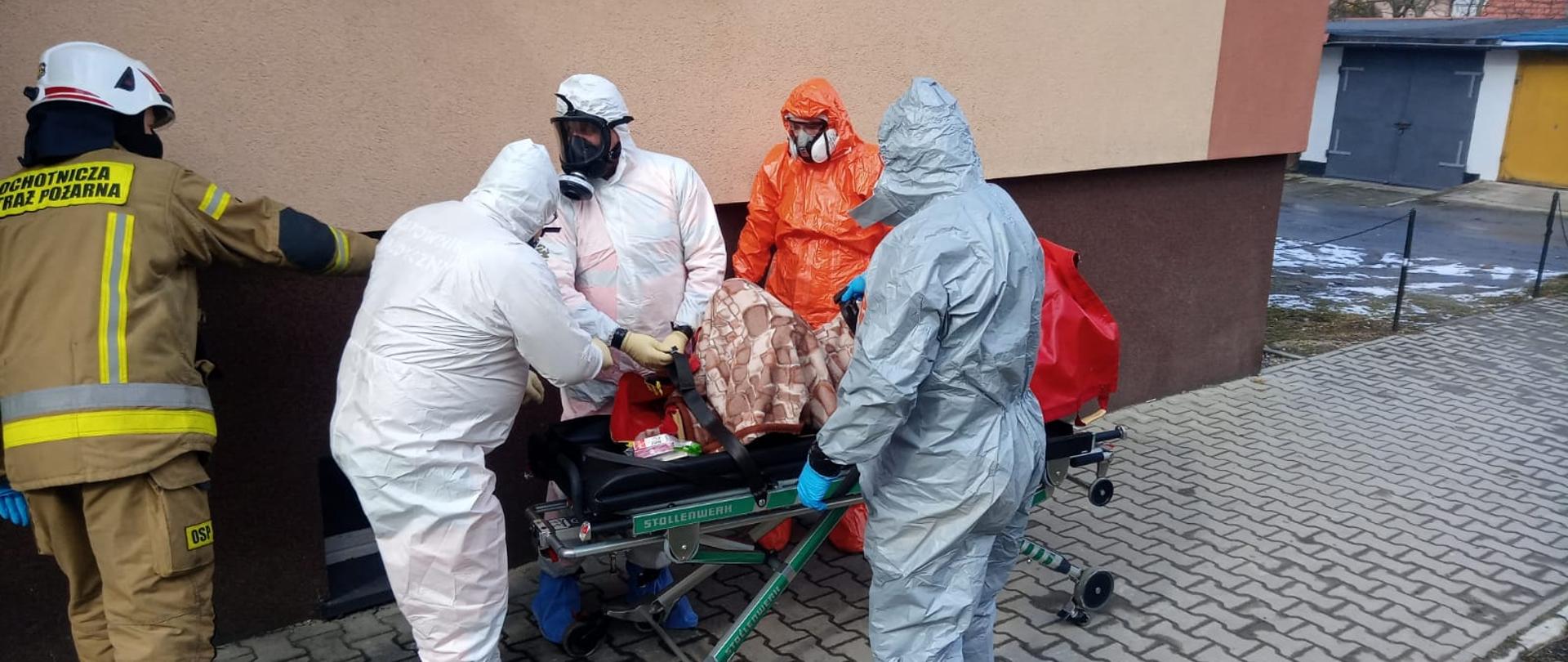 Na głównym planie czterech ratowników Ochotniczej Straży Pożarnej w ubraniu ochronnym. przy pacjencie. Po lewej stronie druh Ochotniczej Straży Pożarnej w ubraniu specjalnym.