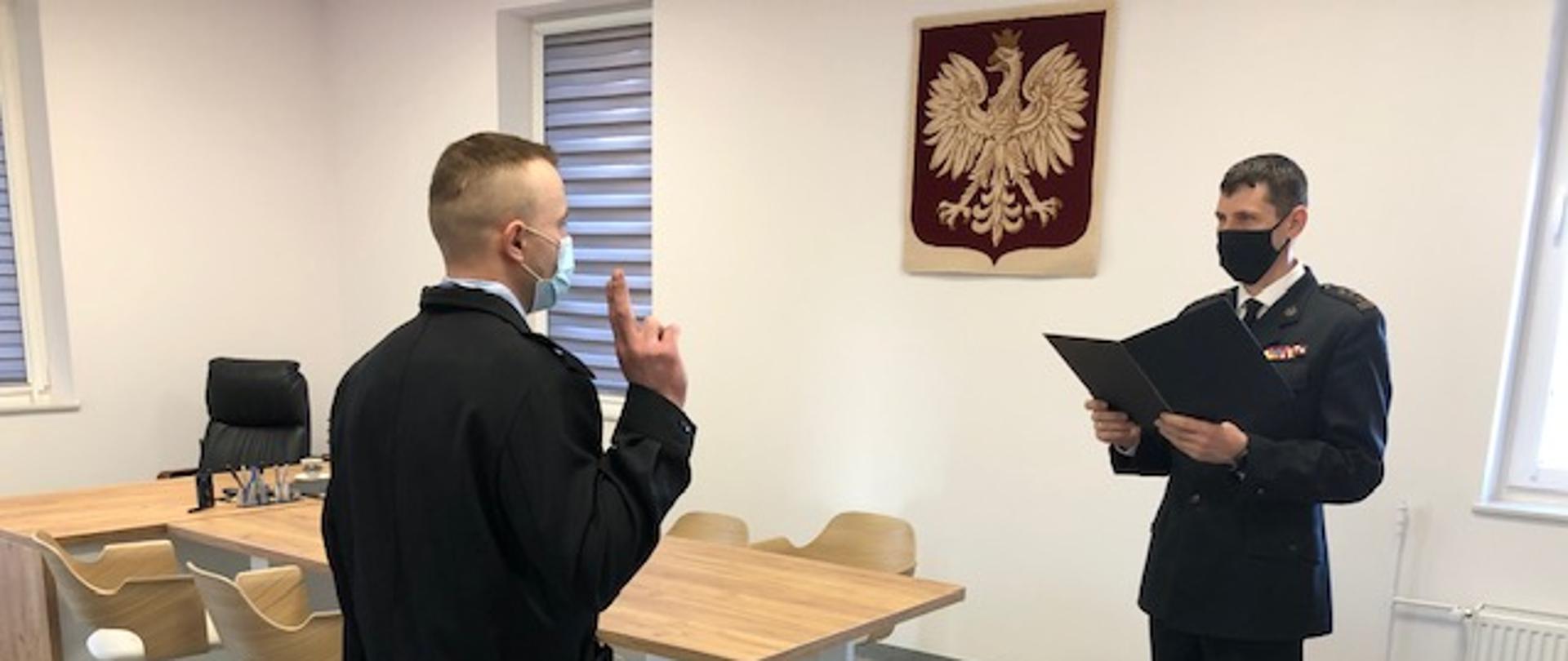 Ślubowanie nowozatrudnionego Strażaka - na tle Godła Rzeczypospolitej Polskiej stoi Komendant Powiatowy PSP w Polkowicach, przed którym ślubuje Strażak