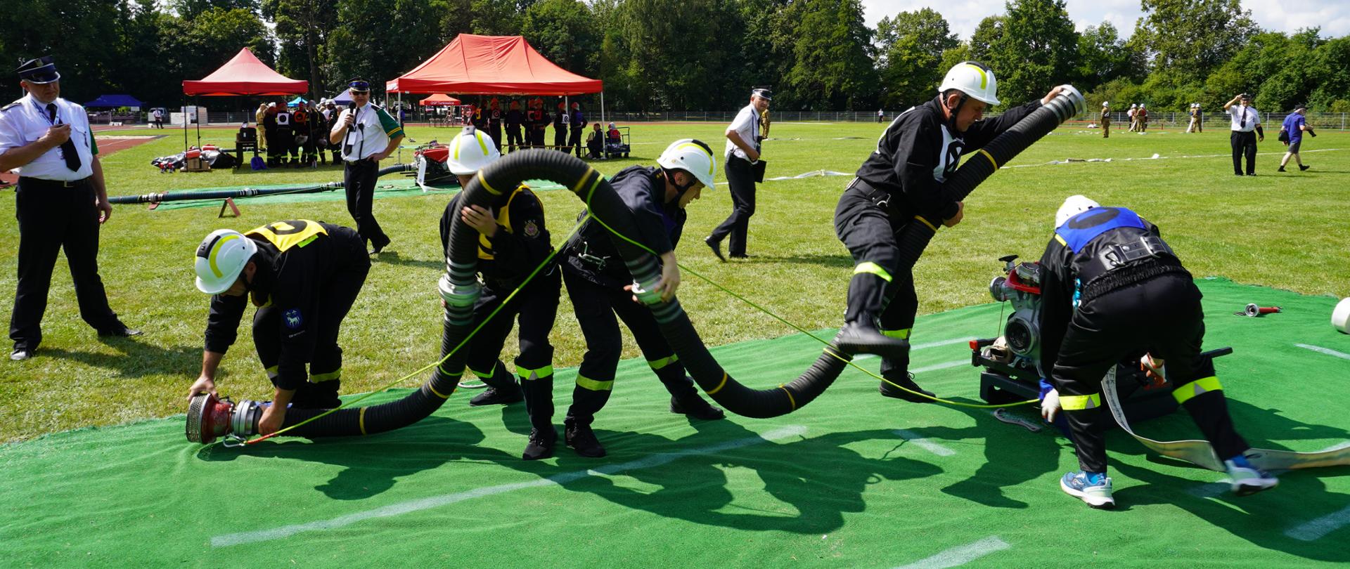 Strażacy w trakcie ćwiczenia sprawiają linię ssawną na zielonej macie. W tle widoczni są sędziowie oceniający wykonanie zadania. 