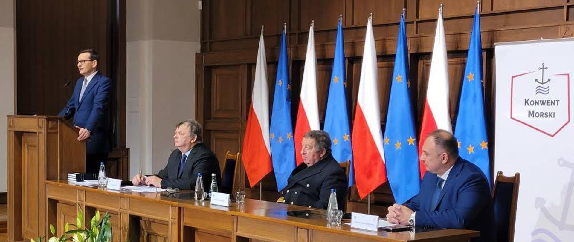 Premier Polski stoi przed mównicą i przemawia za stołem siedzi trzech mężczyzn za nimi są flagi Unii Europejskiej oraz Polski.