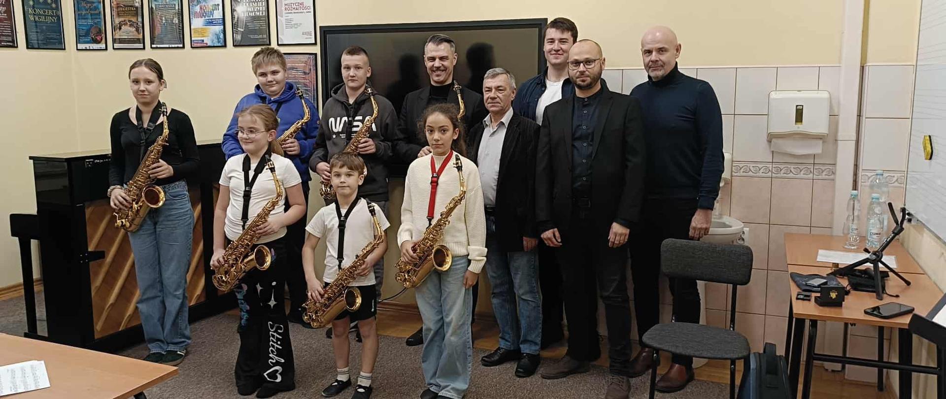 Zdjęcie grupowe wykonane w jasnej sali lekcyjnej. W rzędzie stoi dziesięć osób: pięciu młodych saksofonistów (czterech chłopców i jedna dziewczynka) trzymających saksofony altowe oraz pięciu dorosłych mężczyzn. Wszyscy stoją przodem do aparatu. W tle widoczny jest duży, czarny ekran telewizyjny, plakaty na ścianie, zegar oraz fragment łazienki/aneksu z umywalką i dozownikiem do mydła po prawej stronie. Na pierwszym planie widoczny jest fragment stołu i krzesła. Atmosfera sugeruje zakończenie lub przerwę w zajęciach.