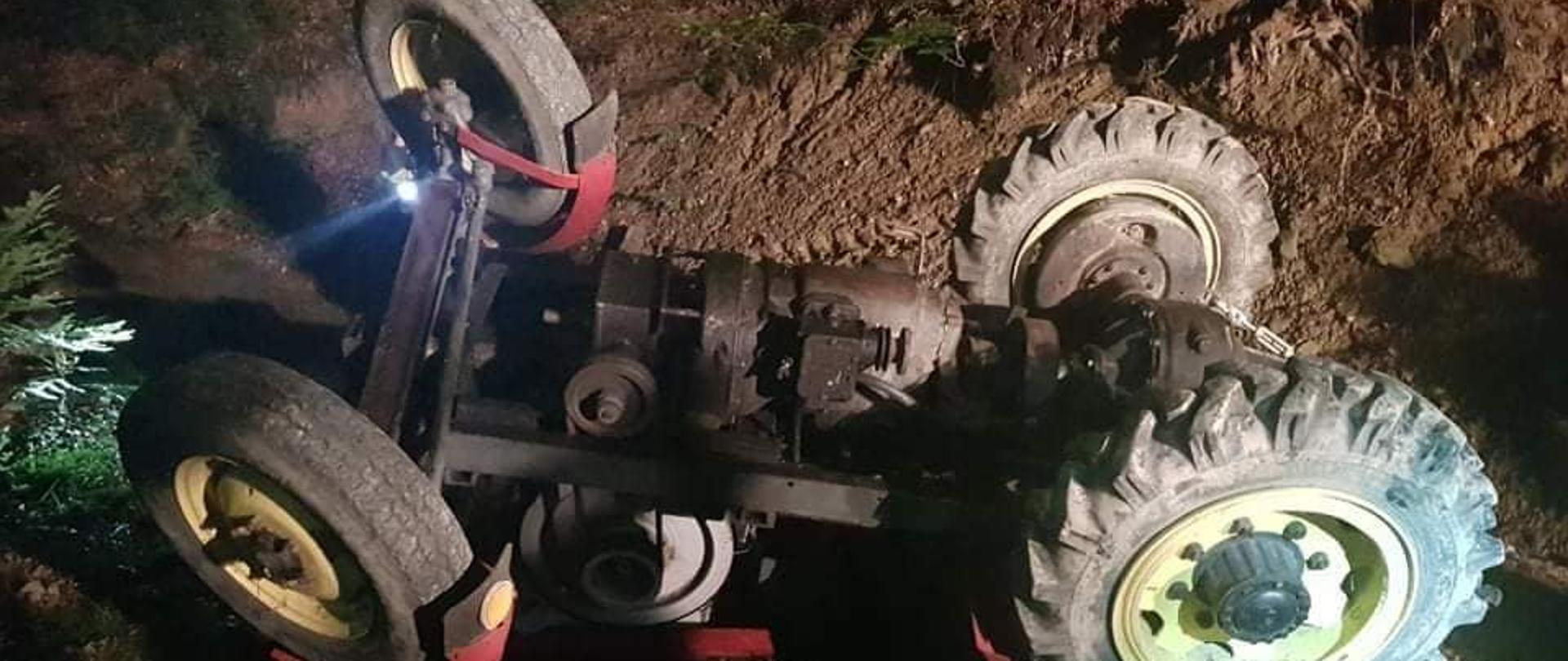 Ciągnik rolniczy leży do góry kołami w lesie.