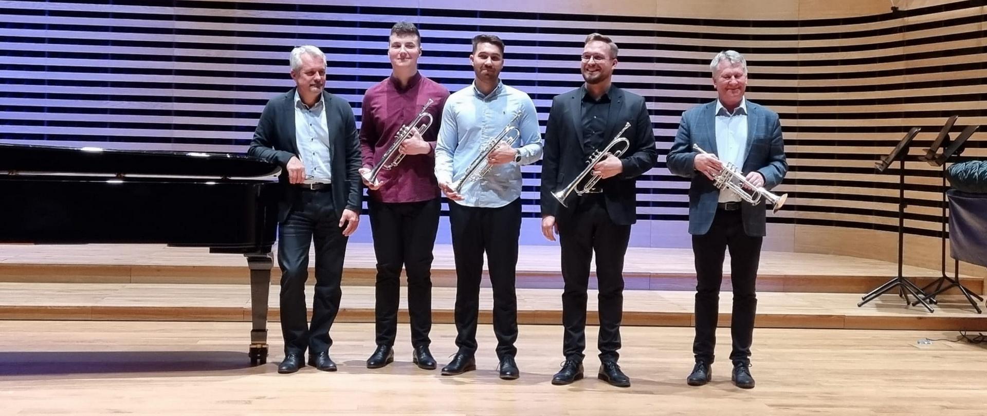 Pięciu mężczyzn stoi w rzędzie na estradzie sali koncertwej, czterech trzyma trąbki w ręce przed sobą, po lewej stronie stoi fortepian