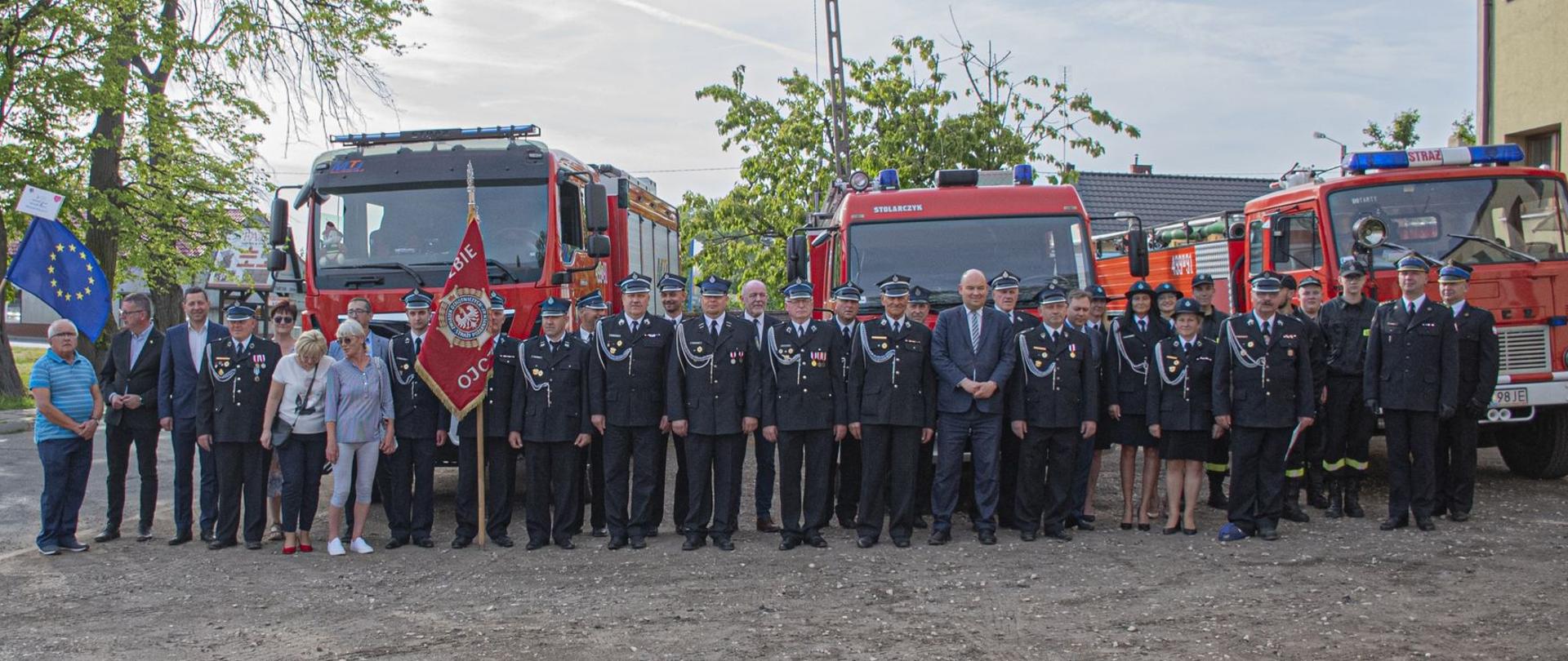 Zdjęcie przedstawia strażaków i zaproszonych gości stojących na tle samochodów strażackich.