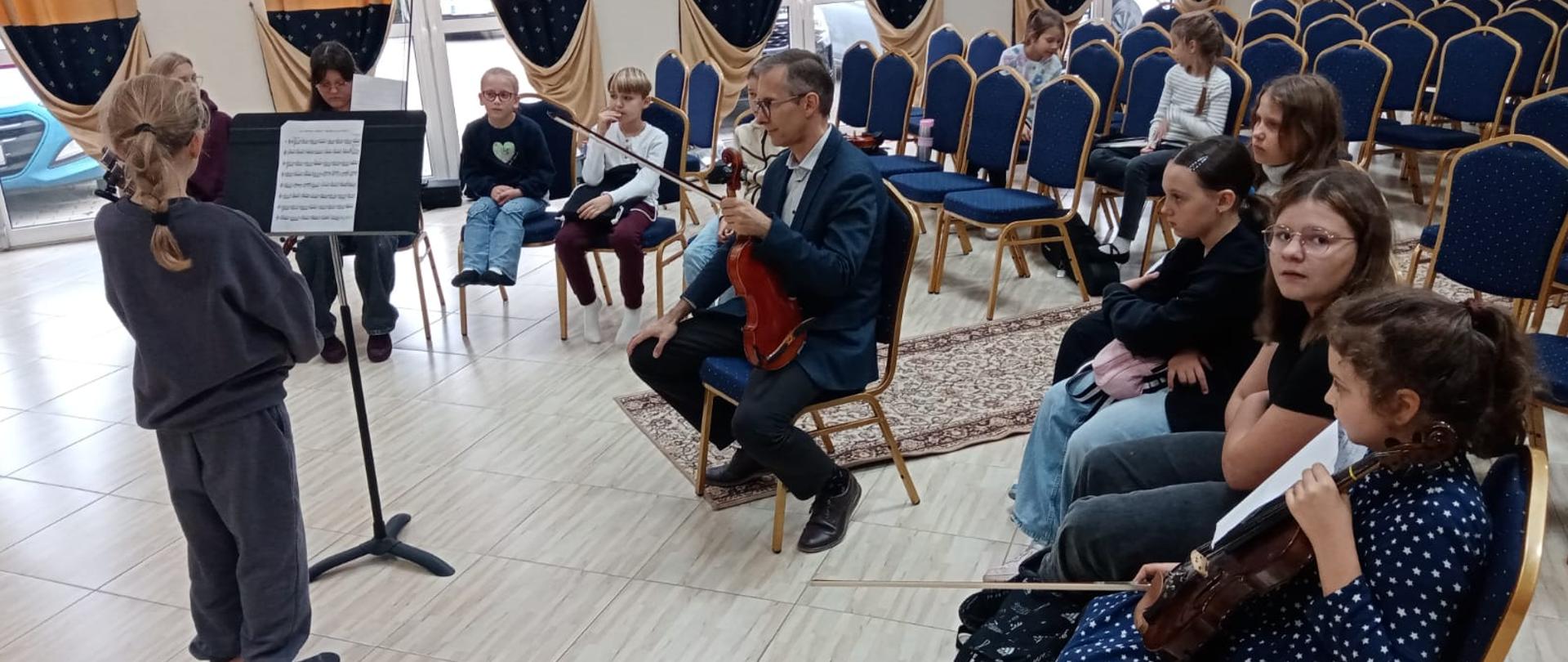 Zdjęcie przedstawia dużą salę z krzesłami ustawionymi w rzędach, oraz częściowo skupionymi wokół mężczyzny ze skrzypcami na których siedzą dzieci obserwujące wydarzenie. Naprzeciw mężczyzny, ubranego w garnitur, noszącego okulary, tyłem stoi dziewczynka o blond włosach splecionych w warkocz, ubrana w dres w ciemnym kolorze, przed nią stoi czarny pulpit z nutami.