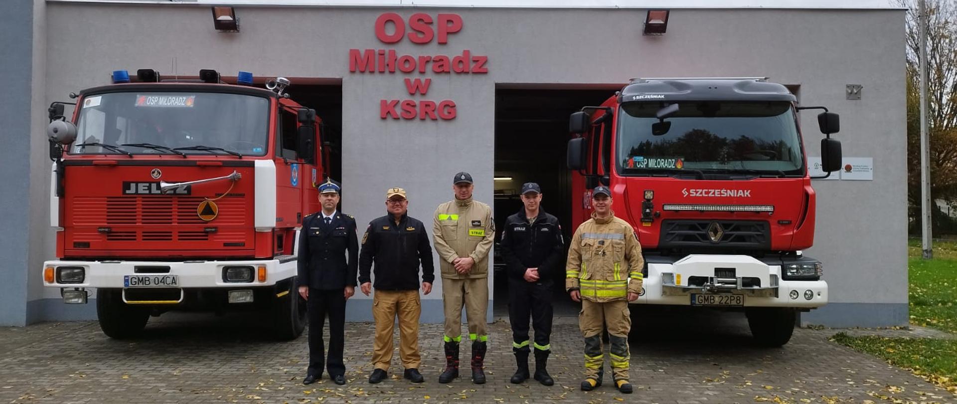 Strażacy Państwowej Straży Pożarnej oraz druhowie Ochotniczej Straży Pożarnej Miłoradz stoją przed remizą obok stoją samochody gaśnicze.