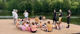 Zdjęcie przedstawia grupę dzieci i dorosłych na drewnianym pomoście nad jeziorem. Na pierwszym planie, po prawej stronie, widać część łabędzia.
Większość osób, w tym liczna grupa dzieci, siedzi na pomoście. Dzieci są ubrane w stroje kąpielowe lub letnie ubrania. Wśród dorosłych stoją trzy osoby w mundurach strażackich lub ratowniczych, prawdopodobnie prowadzące zajęcia lub nadzorujące bezpieczeństwo. Dwóch mężczyzn siedzi dalej na pomoście, tyłem do obiektywu. W tle znajduje się jezioro otoczone gęstym lasem liściastym. Niebo jest częściowo zachmurzone. Scena sugeruje letnie zajęcia rekreacyjne lub edukacyjne nad wodą.