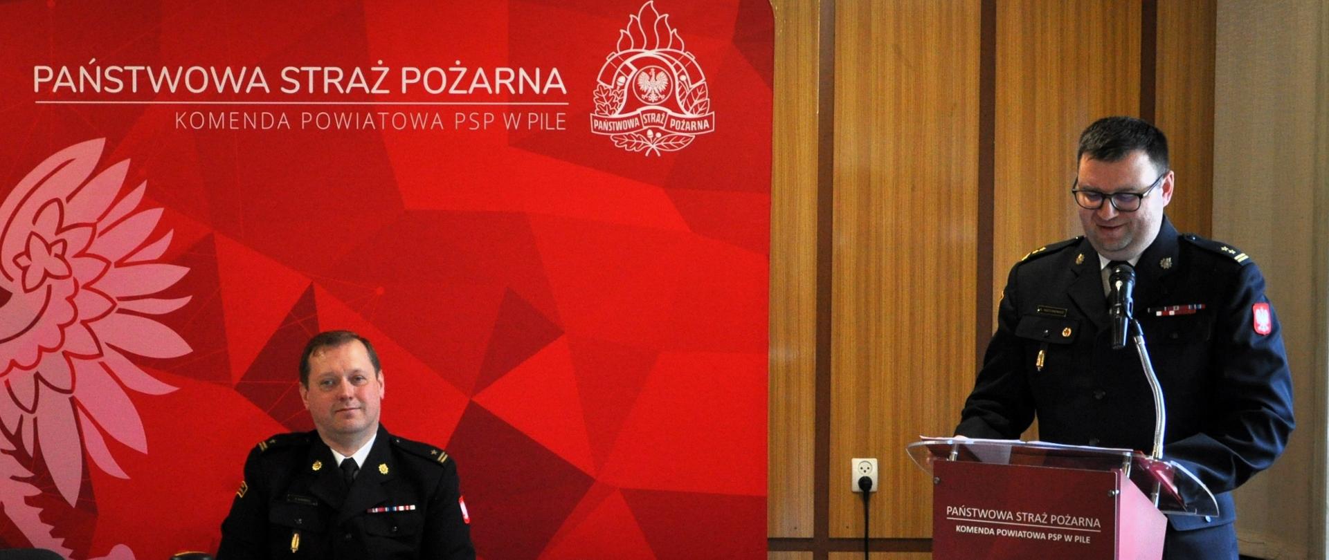 narada strażak siedzi przy stole, obok przy mównicy przemawia strażak, za nimi duża tekstylna czerwona ścianka z orłem 