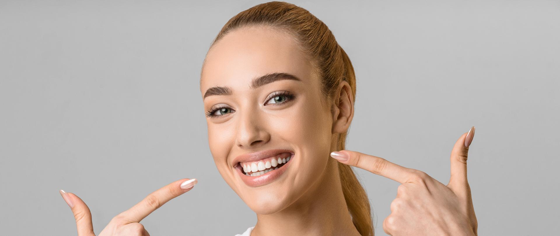 Orthodontic concept. Happy girl showing her beaming white teeth with two forefingers, grey background