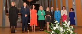 Nauczyciele stoją w jednym rzędzie na scenie sali koncertowej. z przodu przed nimi bukiet z białych kwiatów. Za nimi fortepian a w tle organy