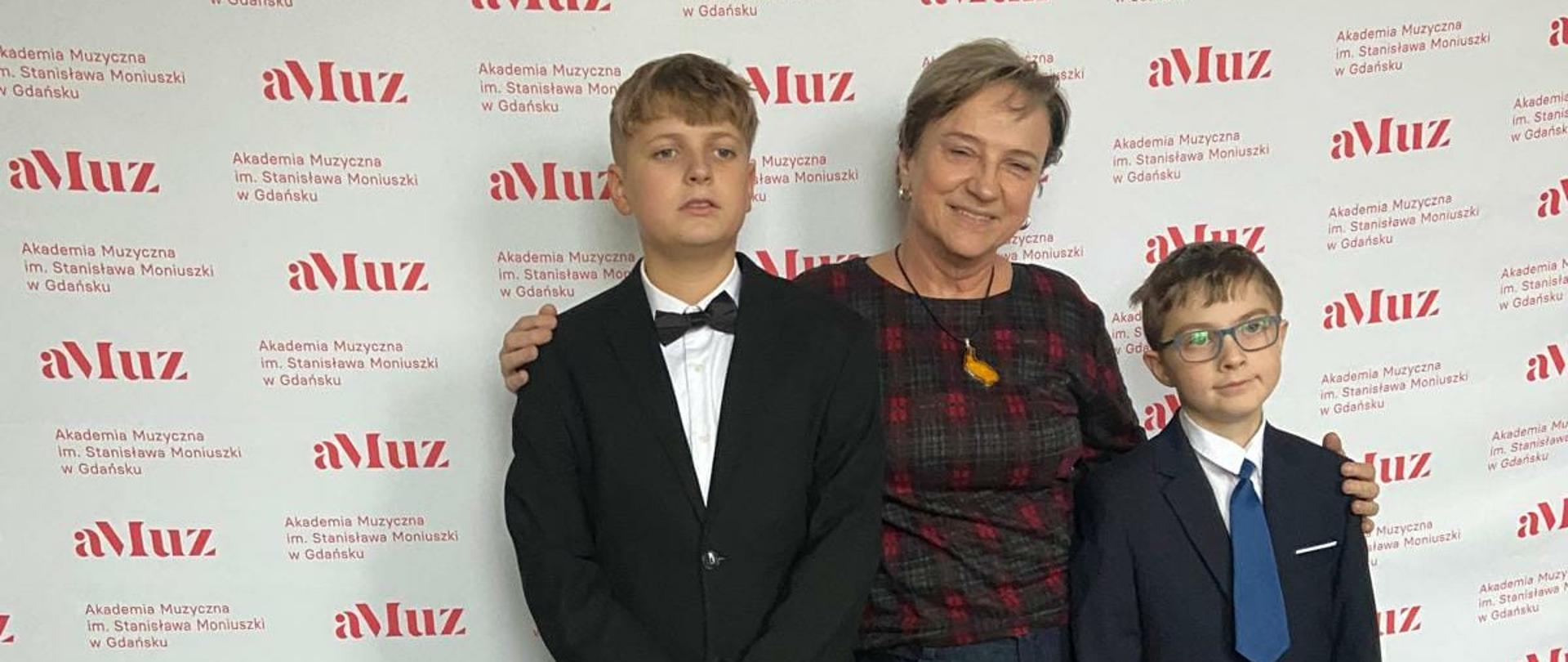 Na tle banera AM w Gdańsku pozuje Elżbieta Lieder wraz z dwójką uczniów w pozycji stojącej. 