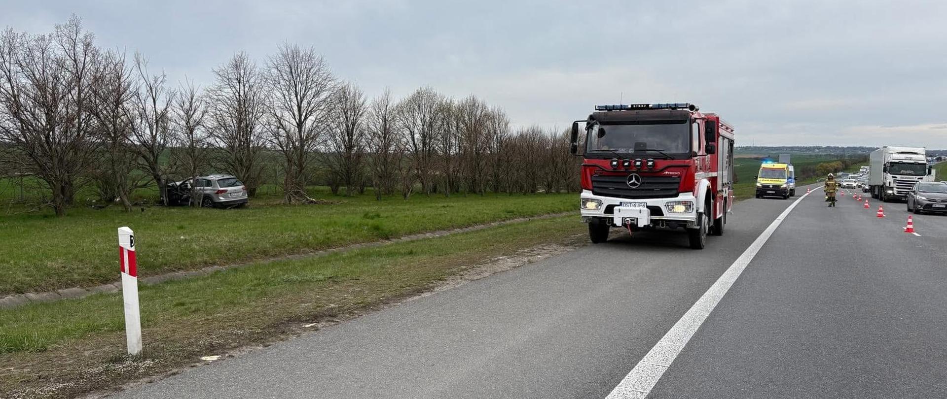 Na zdjęciu znajduje się wrak samochodu osobowego stojący pomiędzy drzewami oraz wóz bojowy straży, karetka pogotowia oraz inne samochody