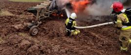 palący się ciągnika rolniczy na polu, za nim dwóch strażaków gaszących pożar przy użyciu linii gaśniczej