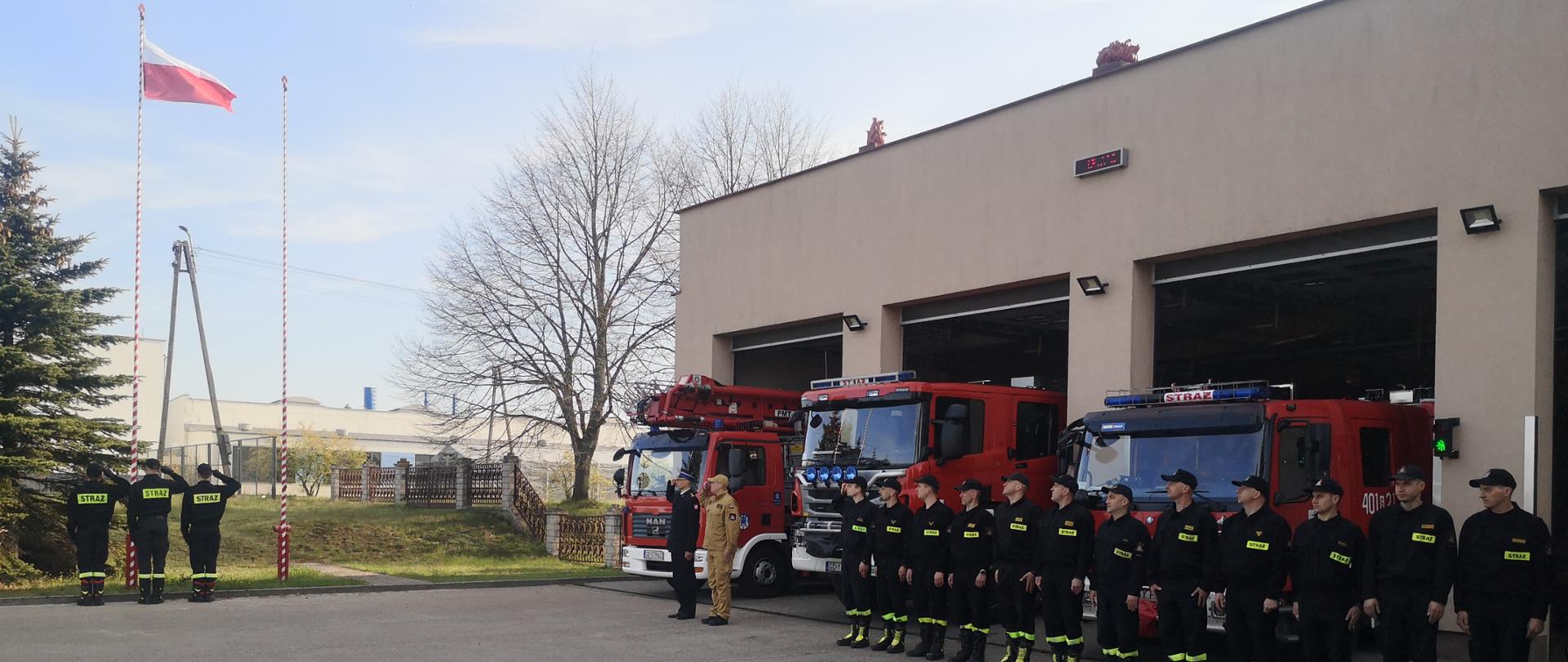 Po lewej stronie w oddali Poczet flagowy salutujący do wciągniętej na masz flagi RP. Po prawej strony wzdłuż garażu JRG ustawieni są w szeregu strażacy stojący na baczność . 2 pierwszych strażaków salutuje do flagi. 