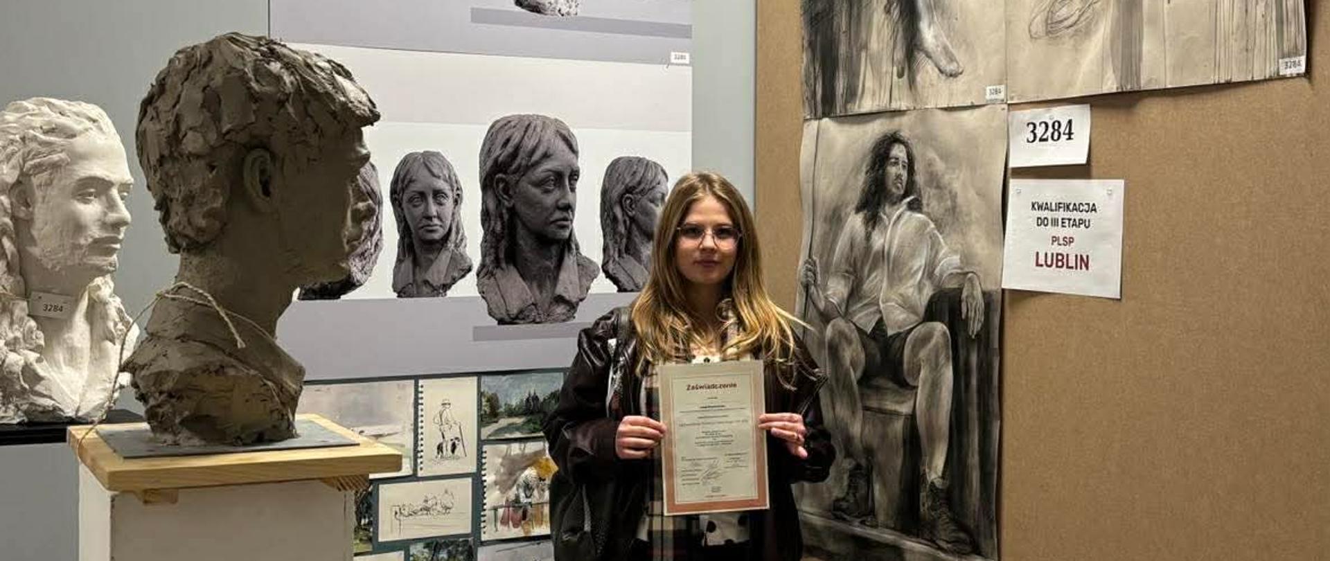 Zdjęcie przedstawia młodą kobietę w okularach, trzymającą dyplom na tle prac plastycznych w PLSP w Lublinie.
Wokół niej widoczne są rzeźby głowy (popiersia) oraz wielkoformatowe rysunki studyjne postaci. Na ścianie obok widnieje kartka z napisem: "Kwalifikacja do III etapu PLSP Lublin.
