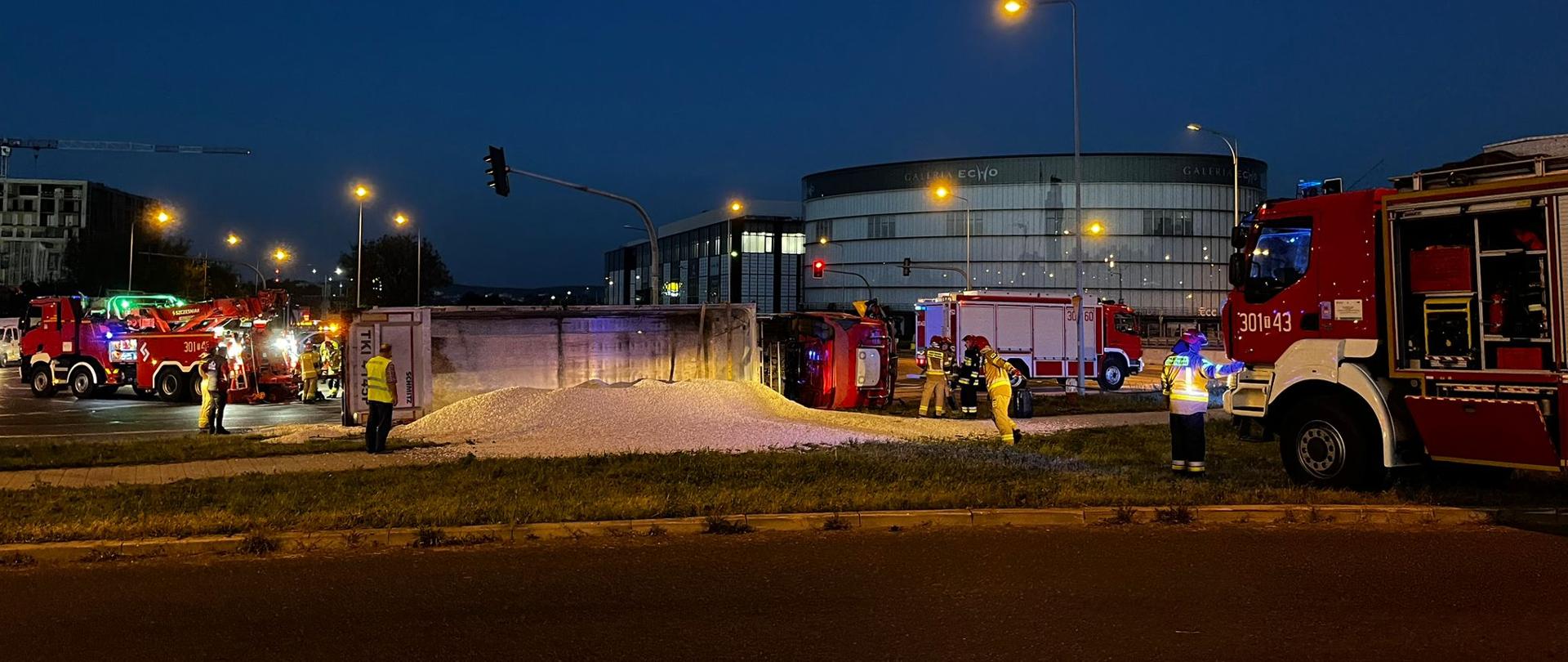 Zdjęcie przedstawia leżąca na boku ciężarówkę z której wysypał się ładunek na pas zieleni. Po prawej stronie i po obu stronach ciągnika stoją trzy wozy strażackie, a przy rozsypanym ładunku widać ratowników. 