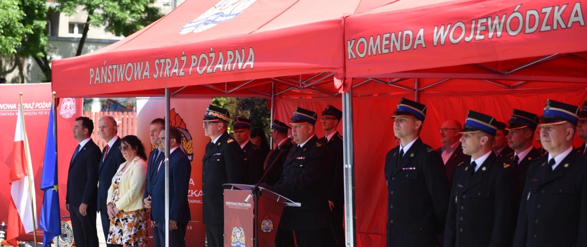 Uczestnicy zbiórki z okazji wręczenia promes na dofinansowanie zakupu samochodów oraz aktów włączenia do KSRG dla jednostek OSP woj. łódzkiego - osoby cywilne i w mundurach galowych PSP stoją pod czerwonym namiotem z napisem PSP, Komenda Wojewódzka