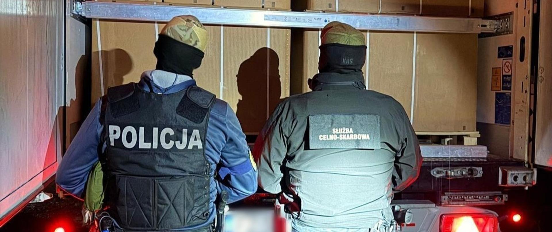 Policjant i funkcjonariusz SCS stoją tyłem przy luku bagażowym samochodu ciężarowego.