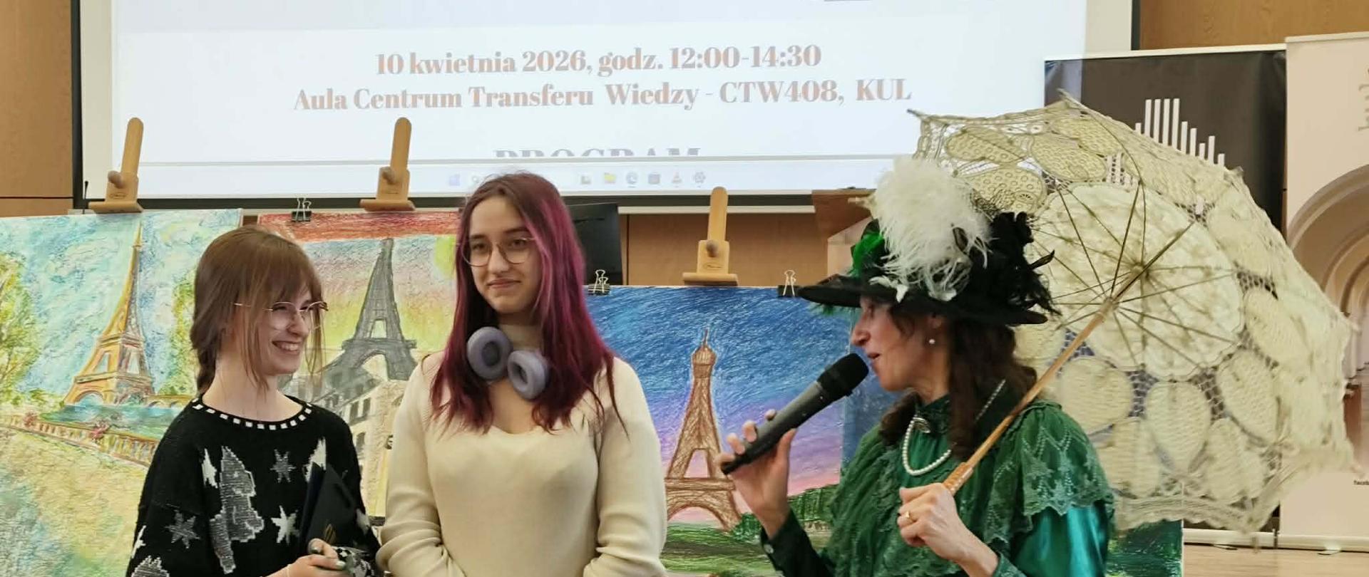 Trzy kobiety na scenie podczas wydarzenia kulturalnego w auli KUL. Po prawej prowadząca w zielonym stroju retro z koronkowa parasolką mówi do mikrofonu, obok niej dwie uśmiechnięte studentki z torbami upominkowymi. W tle obrazy z Wieżą Eiffla.

