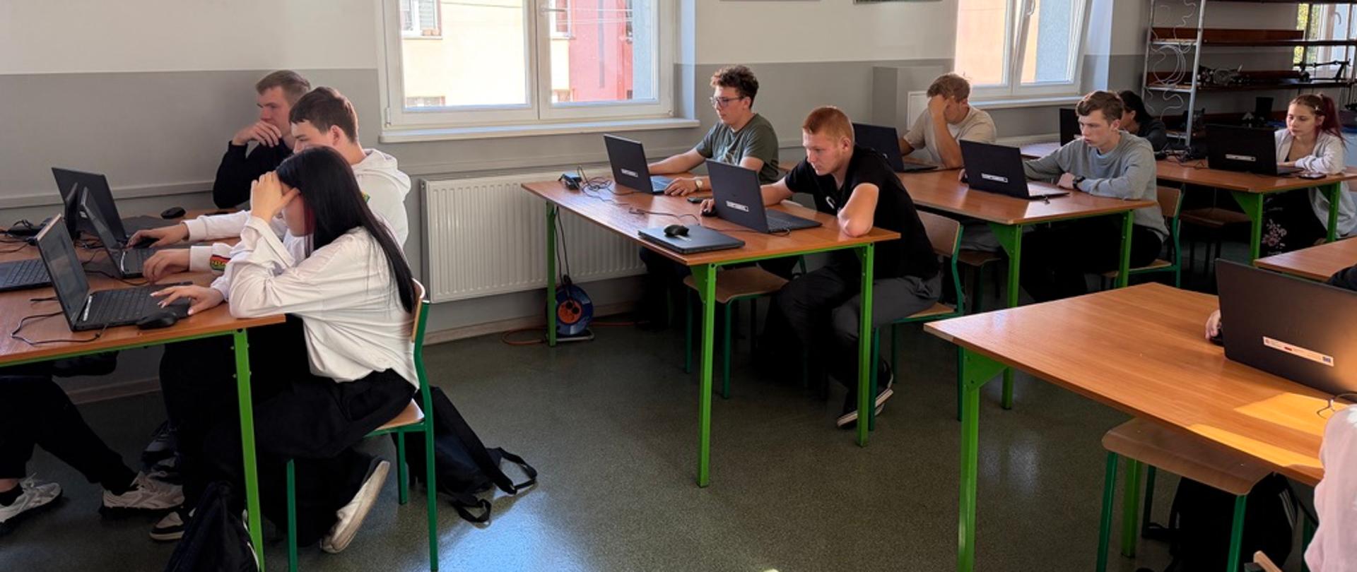 Uczniowie siedzą przy ławkach w sali lekcyjnej i rozwiązują test na laptopach. Na ścianach wiszą zdjęcia maszyn rolniczych pracujących w polu, a po prawej stronie stoi metalowy regał.