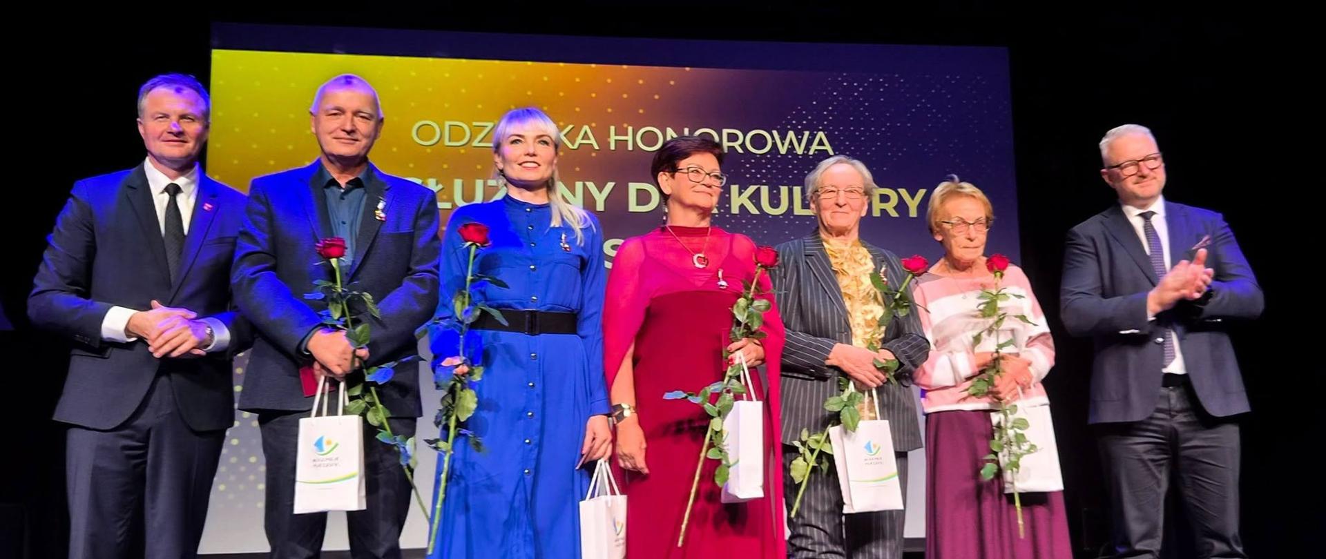 Kilka osób stoi na scenie trzymając róże w ręku 