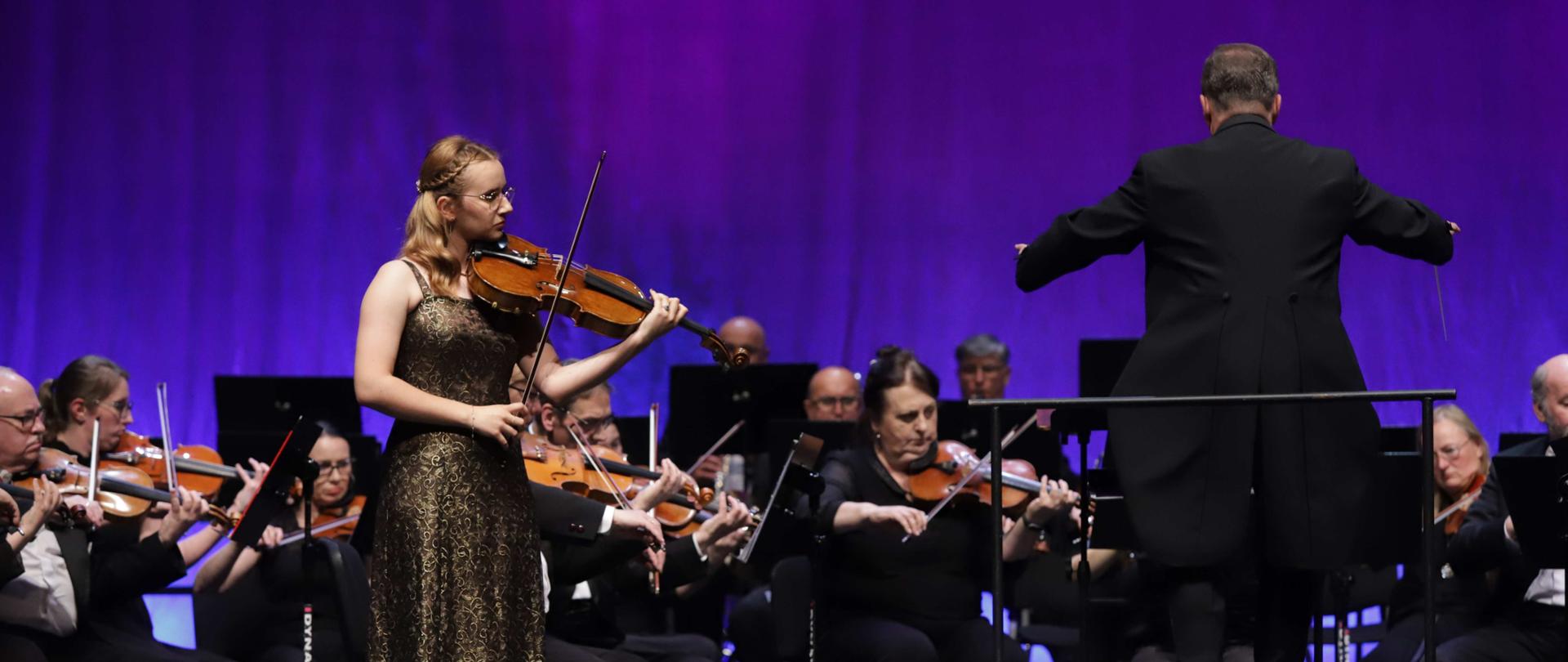 Koncert orkiestry symfonicznej z solistką. Na pierwszym planie młoda altowiolistka Zofia Brych w złoto-czarnej brokatowej sukni wieczorowej wykonuje utwór, stojąc przed orkiestrą. Za nią widoczny jest dyrygent w czarnym garniturze, prowadzący orkiestrę z uniesionymi rękami. Orkiestra składa się z muzyków ubranych w czarne stroje, grających na instrumentach smyczkowych. W tle fioletowe oświetlenie sceniczne tworzy elegancką, koncertową atmosferę. Scena jest profesjonalnie oświetlona, podkreślając uroczysty charakter występu.