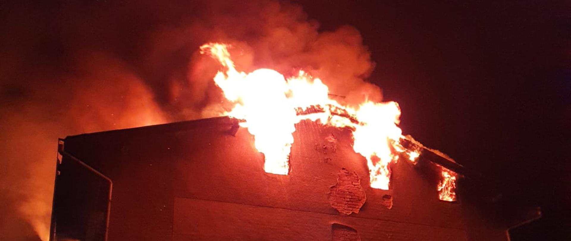 Na zdjęciu widać jedną ze ścian budynku, z okien na poddaszu wydobywają się płomienie i dym.