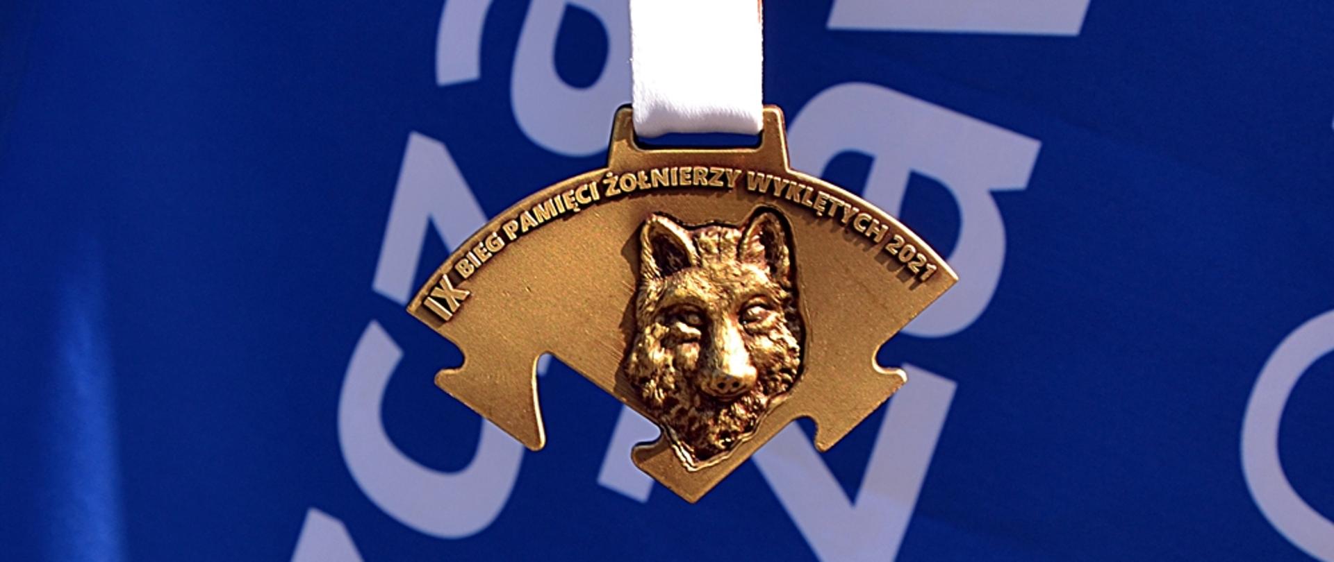 Na zdjęciu widać złoty medal z twarzą wilka i napisem IX bieg pamięci żołnierzy wyklętych 2021 na niebieskim tle z białymi napisami. 
