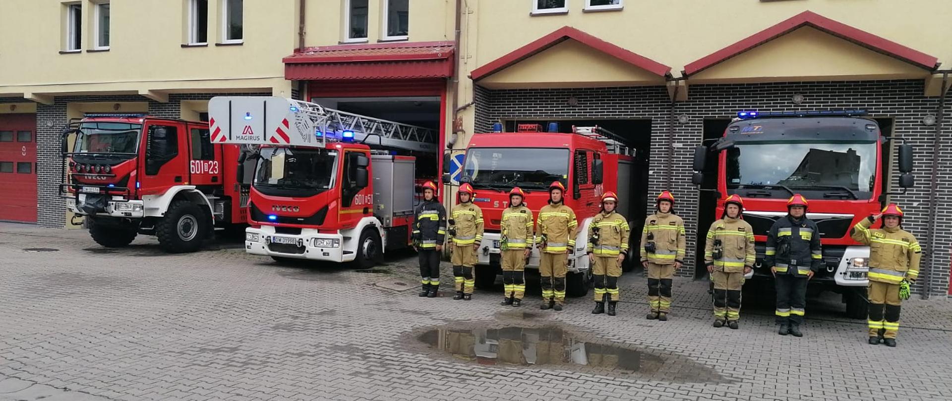 Cztery pojazdy ratowniczo-gaśnicze przed bramami wyjazdowymi z włączonymi synalami. Przed samochodami w szeregu stoi 9 funkcjonariuszy PSP ubranych w umundurowanie specjalne i hełmy. Dowódca zmiany salutuje.