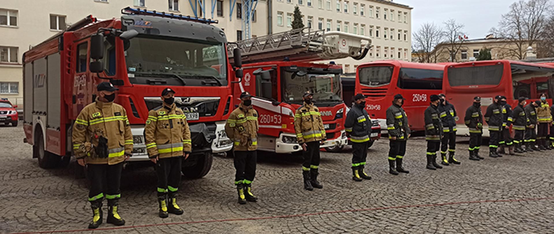 Zdjęcie wykonane na tle budynków. Na pierwszym planie znajduje się strażacy, którzy stoją w szeregu przed samochodami.