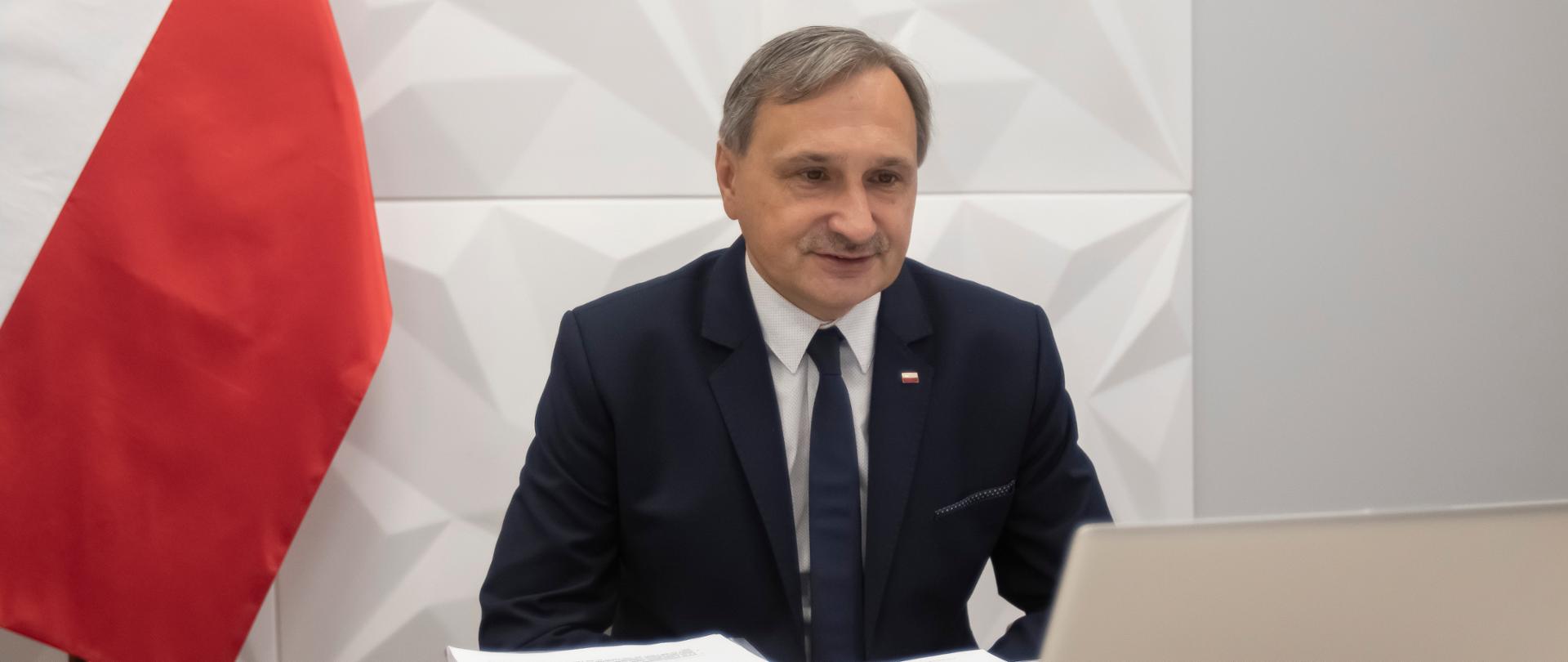 Wiceminister edukacji siedzi przy komputerze. Za nim po lewej stronie flaga biało-czerwona. 