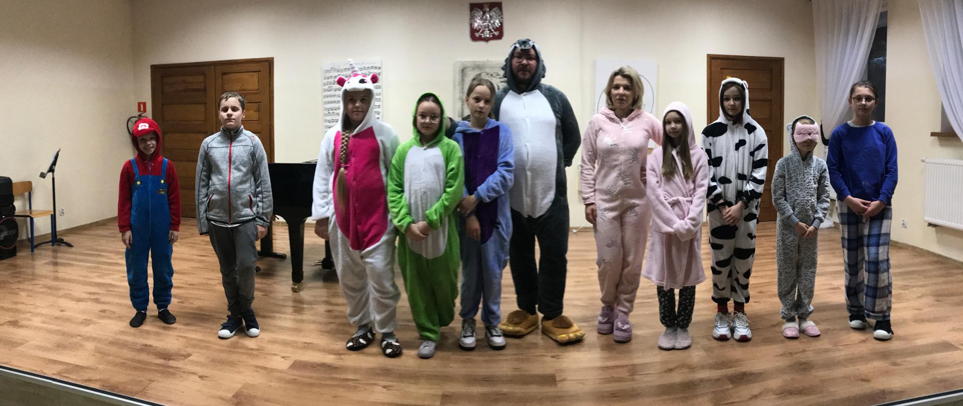 Uczniowie stoją na scenie w szeregu przebrani w piżamy, wraz z nimi nauczyciele Piotr Wilk w piżamie wilka oraz Ewelina Chrzanowska w piżamie jednorożca. 
