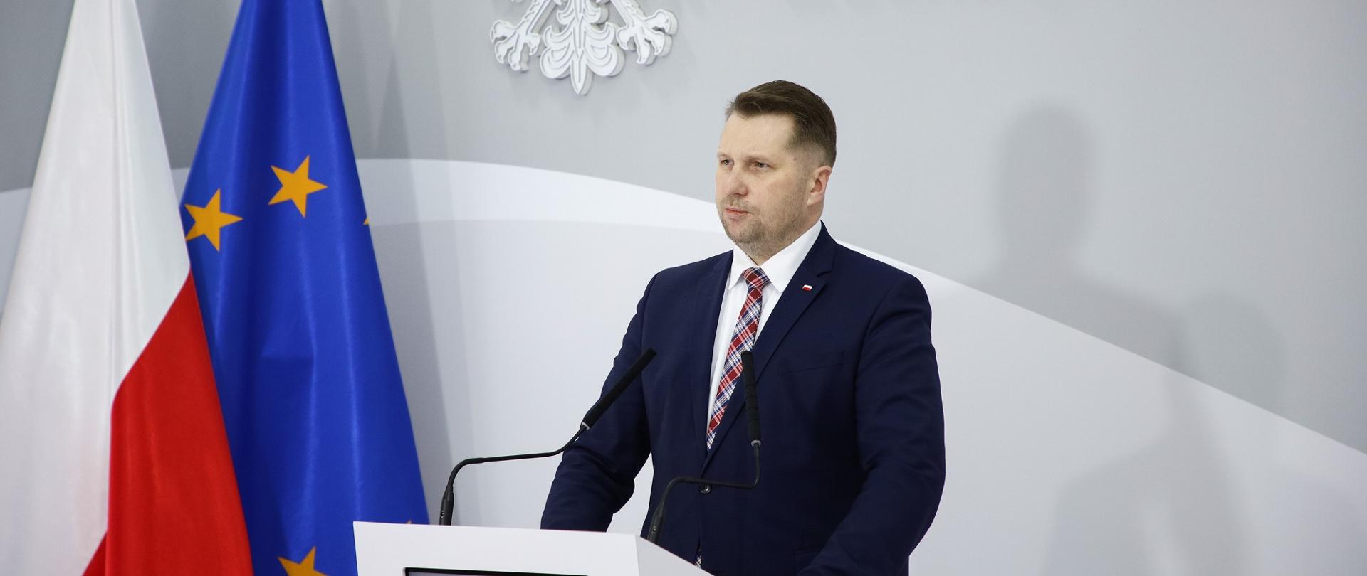 Minister Czarnek przemawia do mikrofonu, za nim flagi Polski i UE.