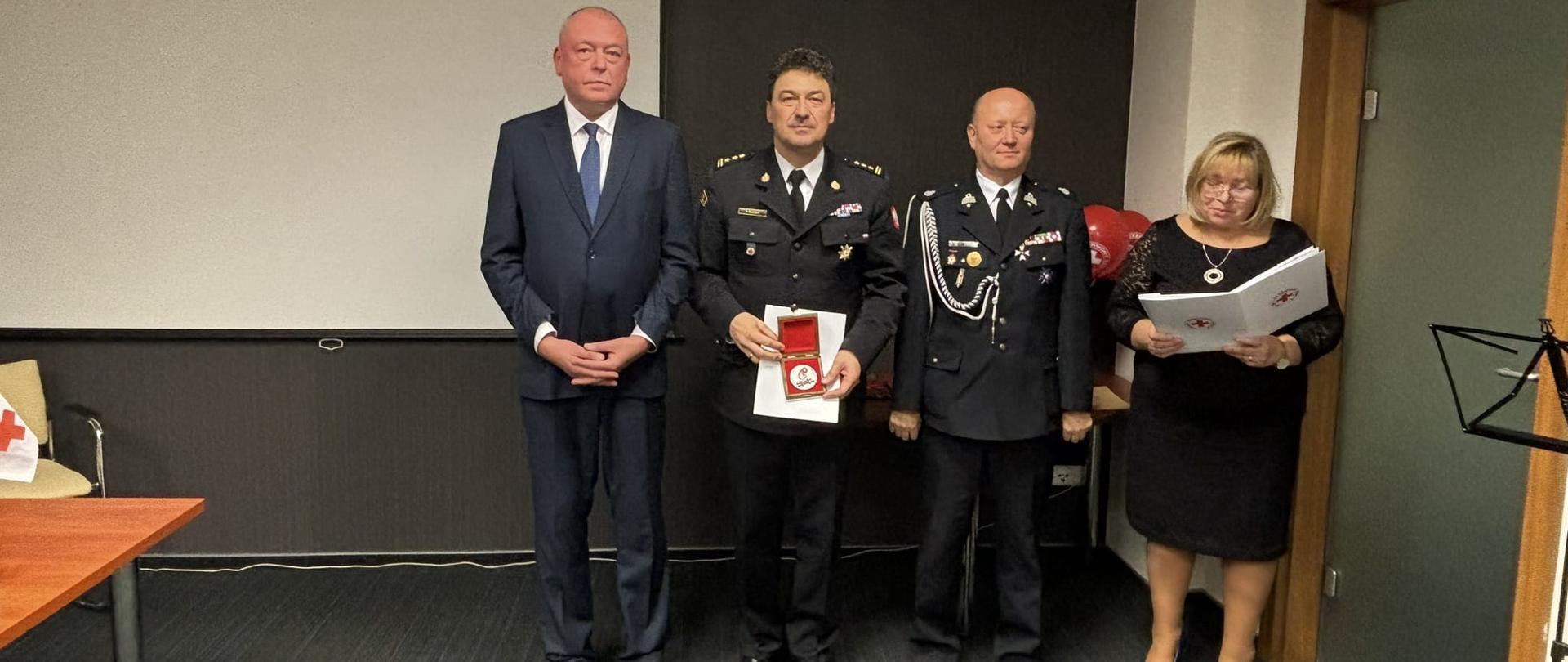 Komendant Centralnej Szkoły PSP trzymający w dłoni otrzymany okolicznościowy medal, po jego prawej stronie stojący mężczyzna po lewej stojący druh OSP oraz kobieta trzymająca w dłoniach białe teczki