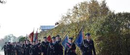 Pododdział strażaków ubranych w umundurowanie galowe maszeruje ulicą. Na czele dowódca pododdziału, następnie osiem pocztów sztandarowych, za nimi pozostali strażacy. 
