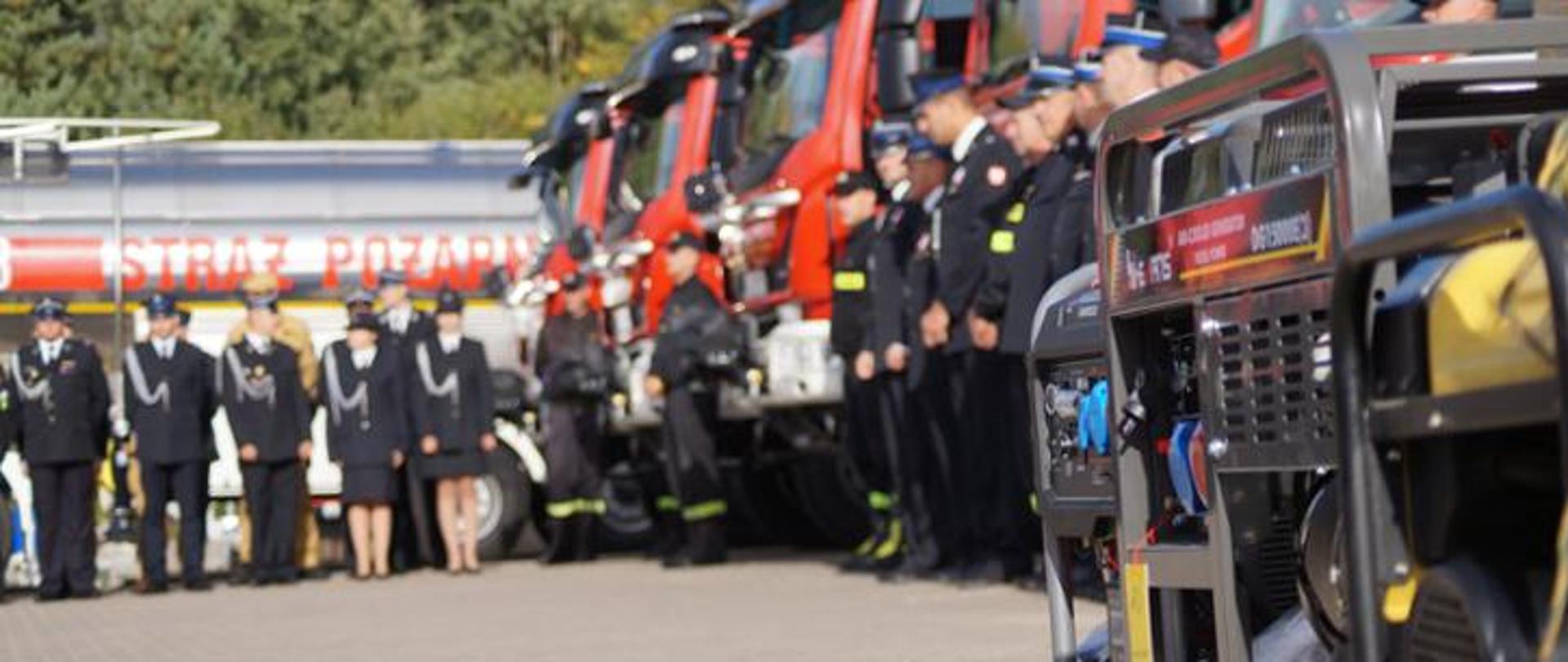 Na tle samochodów pożarniczych stoją strażacy w mundurach.