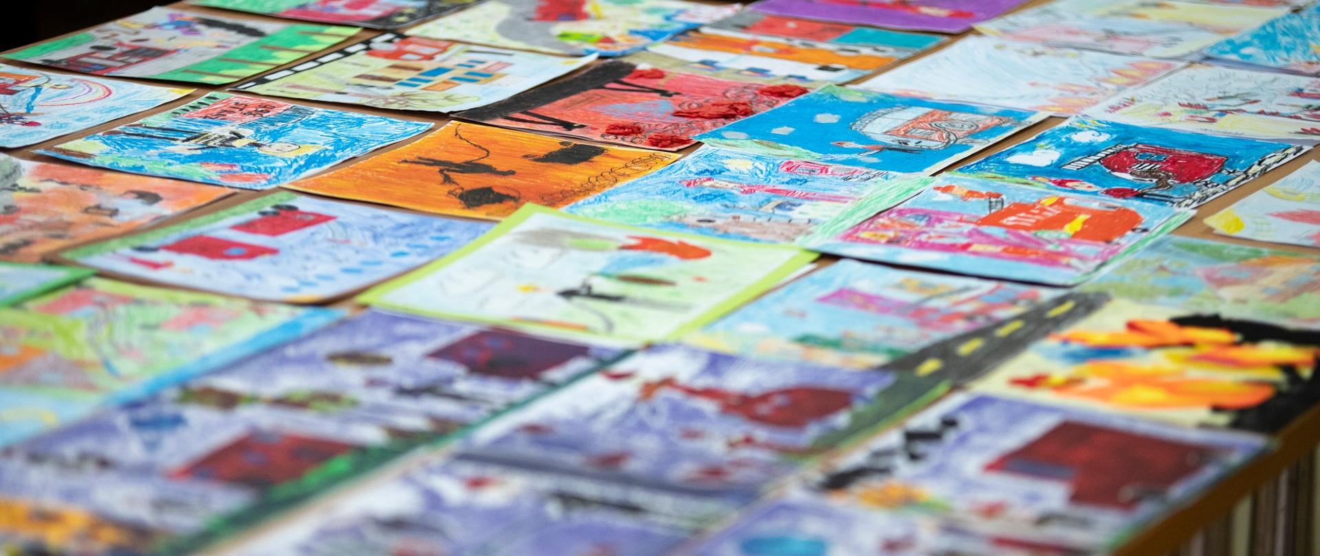 Zdjęcie przedstawia rozłożone na stole prace konkursowe wykonane na kartkach papieru przedstawiające działalność Państwowej Straży Pożarnej 