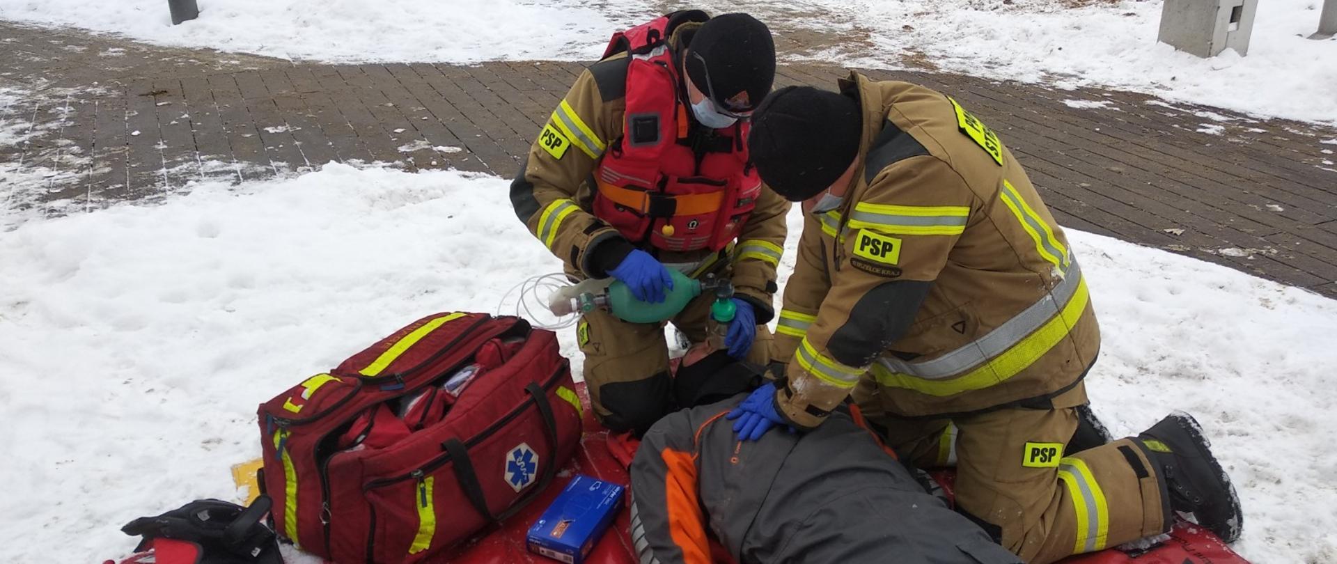 Zdjęcie przedstawia moment udzielania pomocy wyciągniętemu z wody poszkodowanemu. Na czerwonej płachcie rozłożonej na śniegu leży strażak w suchym kombinezonie, pozoruje poszkodowanego nieprzytomnego, po prawej stronie klęczy jeden ratownik, prowadzi ucisk klatki piersiowej poszkodowanego, drugi ratownik u wezgłowia prowadzi sztuczną wentylację z użyciem worka samorozprężalnego, obok na płachcie leży otwarta torba ratownictwa medycznego PSP R1, ratownicy udzielający pomocy ubrani są w ubrania specjalne, mają założone maseczki chirurgiczne na twarzy, w tle widać część skweru przy plaży nad jeziorem Górnym w Strzelcach Krajeńskich. Fotografia wykonana od strony nóg poszkodowanego.