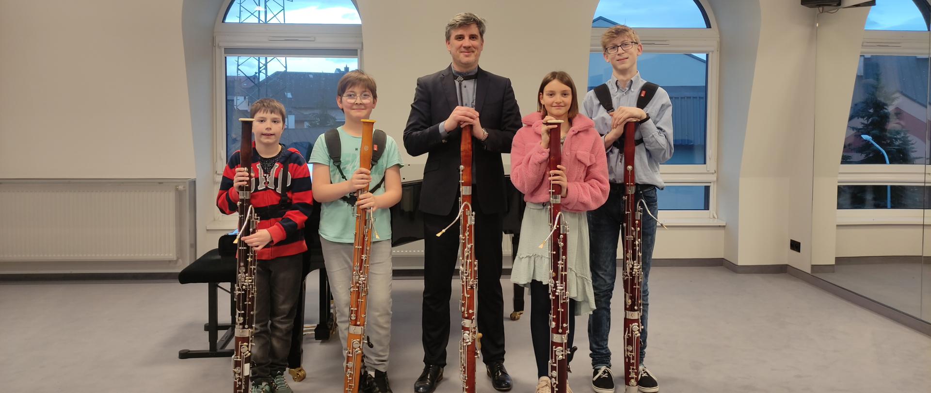 Uczniowie pozują do zdjęcia podczas warsztatów fagotowych. W środku prowadzący warsztaty po jego prawej i lewej stronie uczniowie biorący czynny udział w warsztatach fagotowych. Wszyscy trzymają fagoty ułożono pionowo z oparciem na podłodze. Za nimi fortepian. Po prawej stronie lustra, po lewej grzejnik, w tle okna